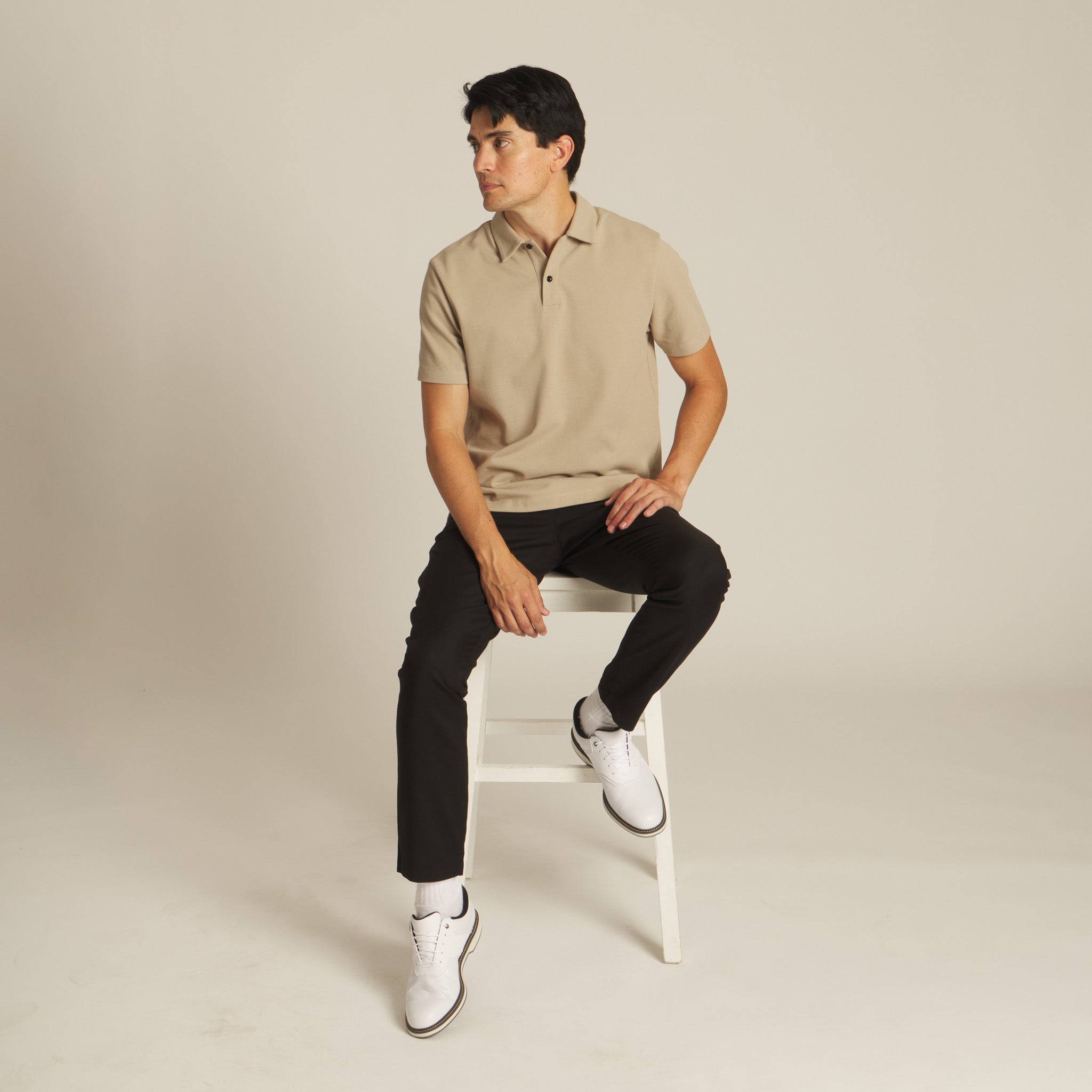 Man sitting on a white stool wearing a beige Mentfore polo shirt and black pants against a beige background